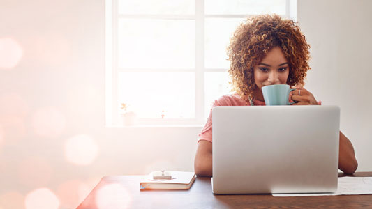 A woman in a bright sunny room sitting in front of her laptop and happily sipping on coffee