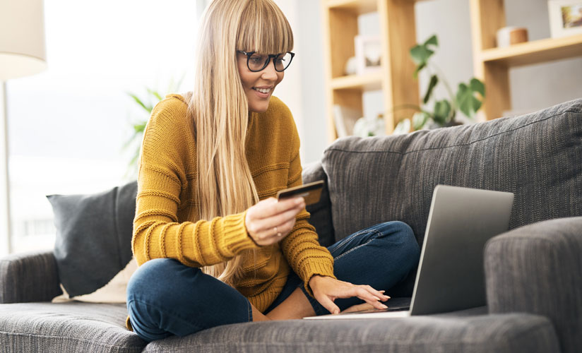 A young blonde woman sitting on the couch in front of her laptop holding a credit card, presumably excited about what she is ordering online at low cost