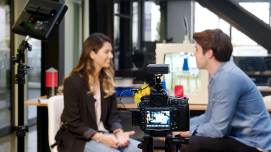 A man and a woman sitting across from each other having an interview, the shot is taken from behind the camera that's filming them